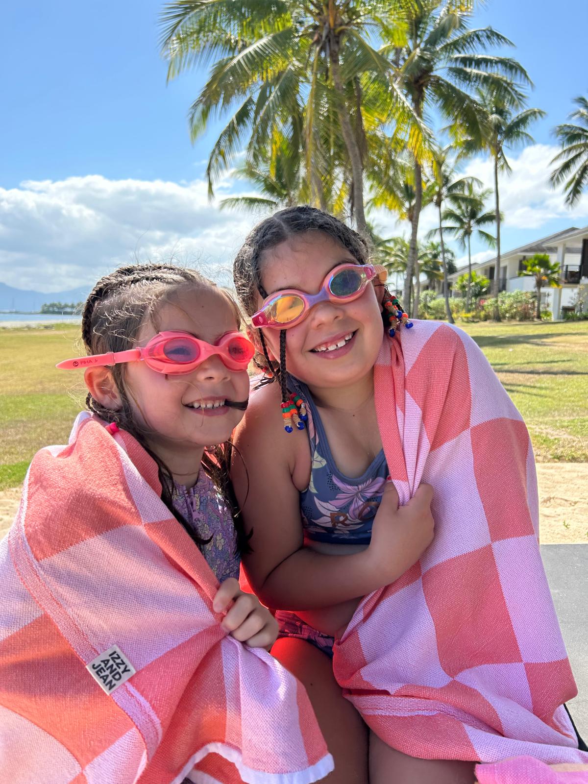 Two children wrapped in a pink towel on a beach with palm trees in the background