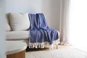 The Riveria Dark Blue throw blanket with tassels draped over a beige sofa in a bright room.