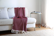 Burgandy throw blanket with white tassels strewn over the back and seat of a white sofa in a living room.