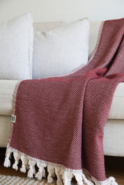 A burgundy throw blanket made from 100% cotton, with a herringbone pattern and fringed edges on a white background on a white couch in a living room.
