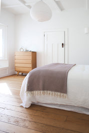 Bed with a brown riveria throw blanket in a room with wooden flooring and a dresser.