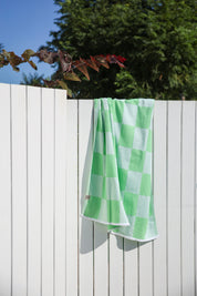 Green and white checkered towel hanging on a wooden fence with trees in the background