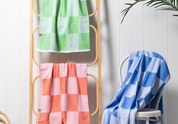 apple green, coral and blue checkered Turkish towels hanging on a ladder shelf and white wooden chair with a wooden white wall behind them and plant in the corner.