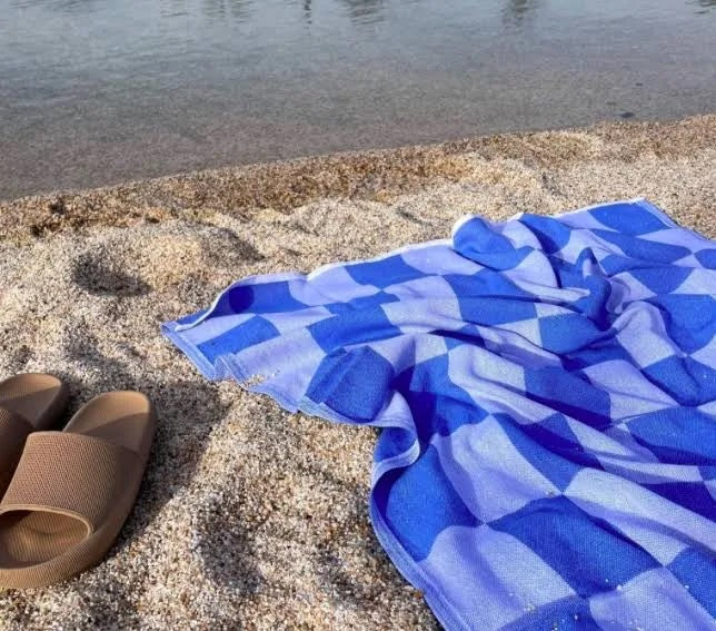 A checkerboard-patterned towel in royal blue, laid out on sand beside a pair of brown sandals, with a body of water in the background.