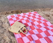 A coral and white checkerboard-patterned Turkish towel laid out on the sand with a beige tote bag on top of it, near a body of water.