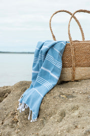 The Aegean Blue Turkish Towel in a weave basket on the sand with the sea in the background