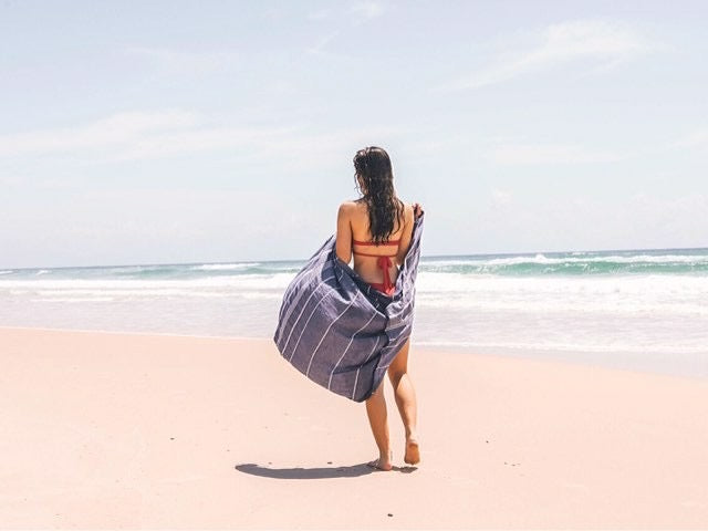 A girl in a red bikini walks on the beach with a navy blue Turkish towel - classic wrapped around her