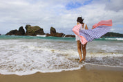 a lady in a white dress playing in the shallow waves on the beach with a blue and coral striped Turkish towel bellowing out behind her.