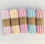 5 rolled stripey Turkish towels in a row, with Izzy and Jean mid paper branded wrappers. Colours start with purple, yellow, pink, ocean and coral and white stripes.
