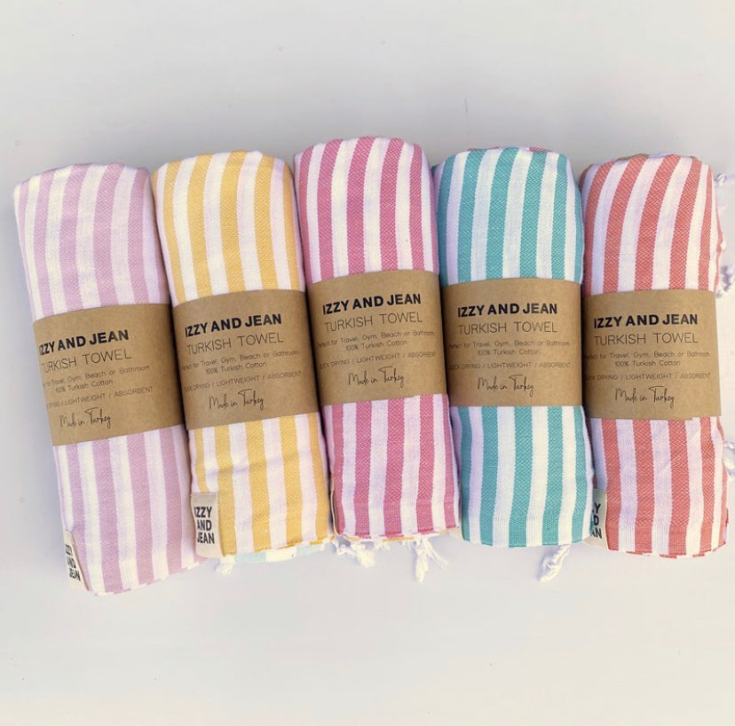 5 rolled stripey Turkish towels in a row, with Izzy and Jean mid paper branded wrappers. Colours start with purple, yellow, pink, ocean and coral and white stripes.