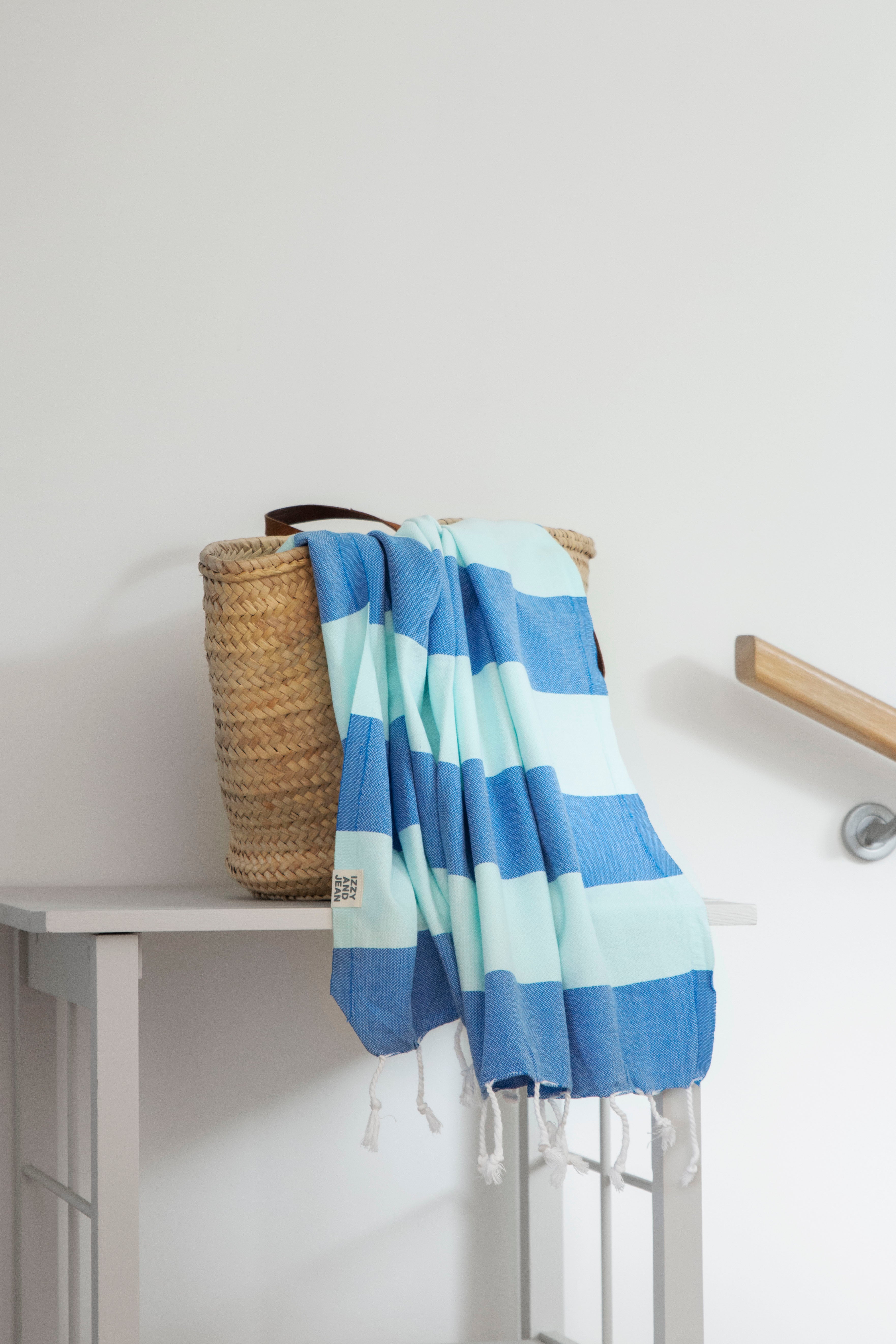 A folded striped towel with bands of blue and mint colours, hangs over a weave basket on a white table