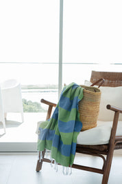 A striped Santorini Turkish towel with navy and olive colours, hangs over the edge of a weave basket on a stylish rattan chair with white pillows