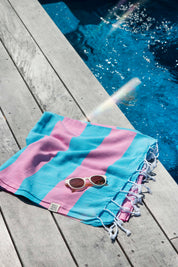A folded Santorini Turkish towel with marine blue and hot pink stripes with white sunglasses on top is placed beside a pool on a wooden deck 
