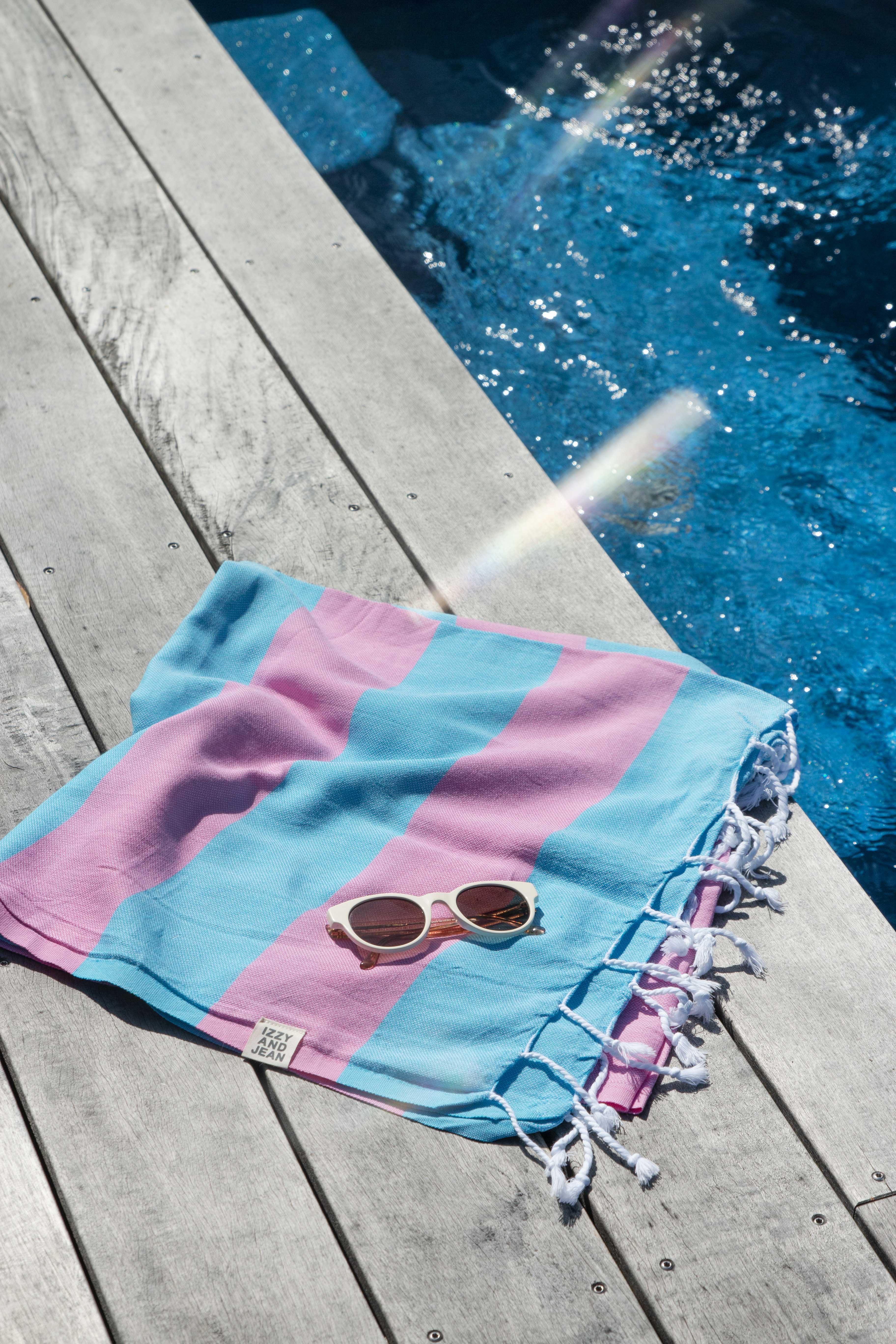 A folded Santorini Turkish towel with marine blue and hot pink stripes with white sunglasses on top is placed beside a pool on a wooden deck 