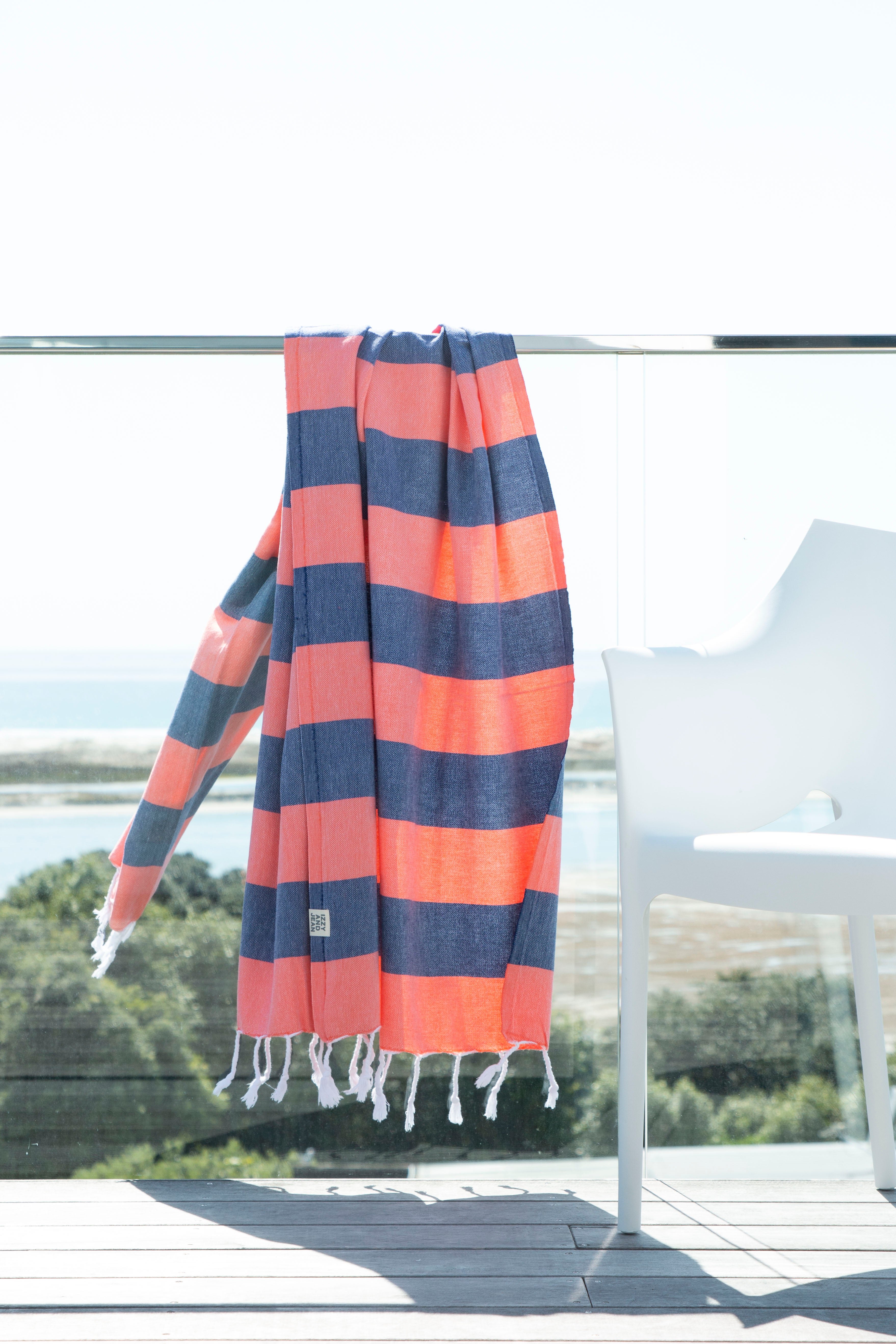 A Santorini Navy and Coral Striped Turkish Towel lying on a glass banister over-lookng a beach