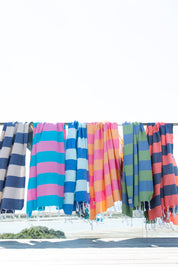 A range of Santorini Striped Turkish Towels on a glass banister overlooking the beach