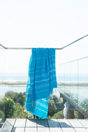 A marine-coloured Classic Turkish towel with white stripes and fringes hangs on a glass banister on a deck overlooking a beautiful ocean vista