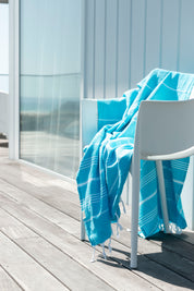 A marine-coloured Classic Turkish towel with white stripes and fringes hangs over a white chair on a wooden deck