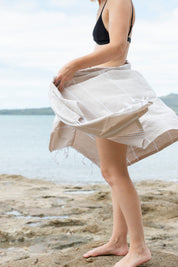 a girl wears a black bikini and stands near the sea wearing the sand-coloured Classic Turkish towel around her wiast.