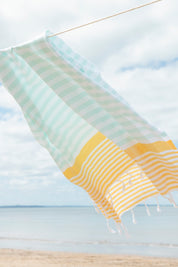 A mint, yellow and white striped Turkish towel blowing in the wind on a washing line with the beach and sand in the background