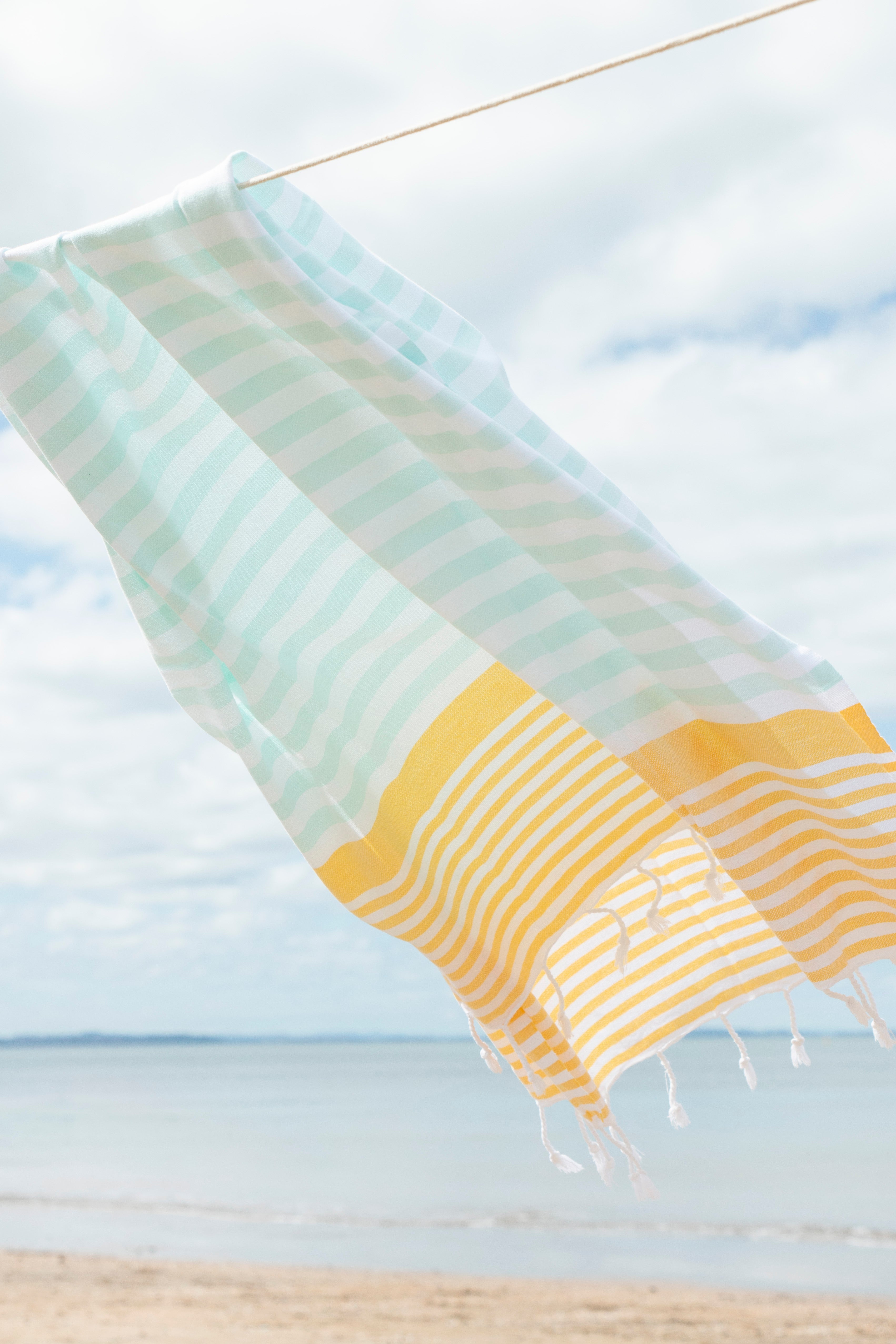 A mint, yellow and white striped Turkish towel blowing in the wind on a washing line with the beach and sand in the background