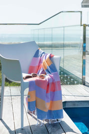 A striped multi-coloured turkish towel in shades of blue, pink, and orange is draped over a white chair, with a pool and balcony in the background.