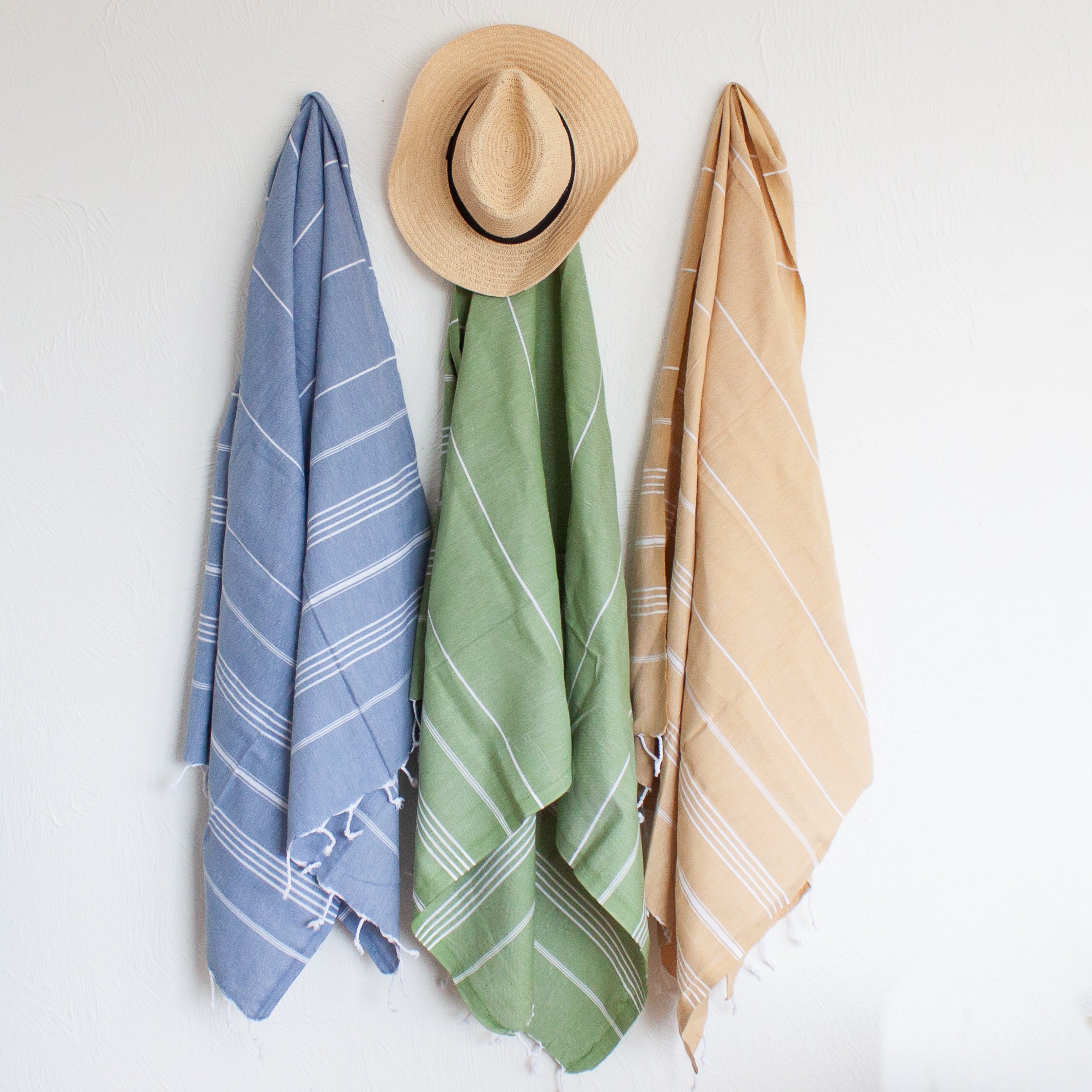 Three Turkish towels with white stripes and fringes on the bottom, hanging on hooks in navy, olive and yellow with a straw hat placed on top of the middle hook