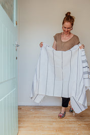A smiling lady holds up a white Turkish towel with black stripes and tassels on the bottom edge inside her white wooden front door with wooden floors